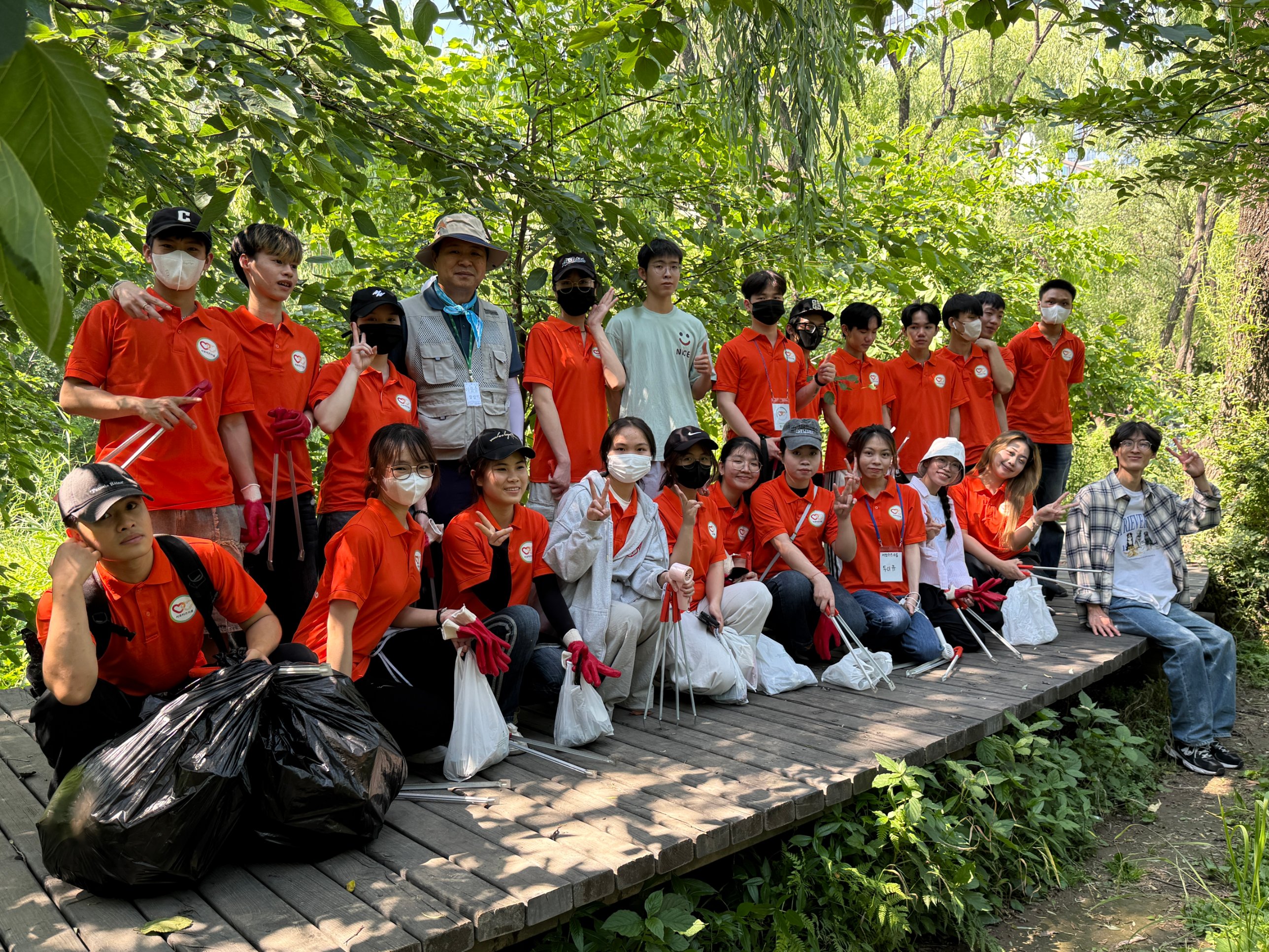 Group of young volunteers in orange polo shirts posing in a leafy wooded setting during a community service outing