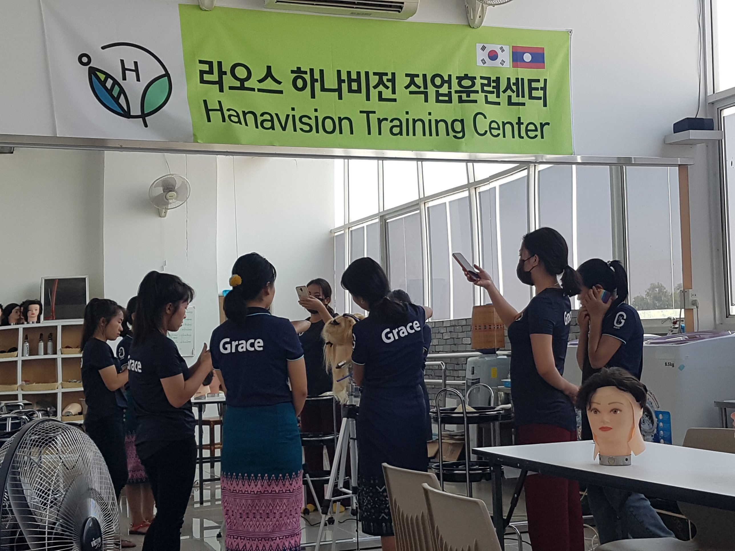 Trainees in Grace Beauty Academy uniforms working with mannequins beneath the HanaVision Training Center banner in Laos