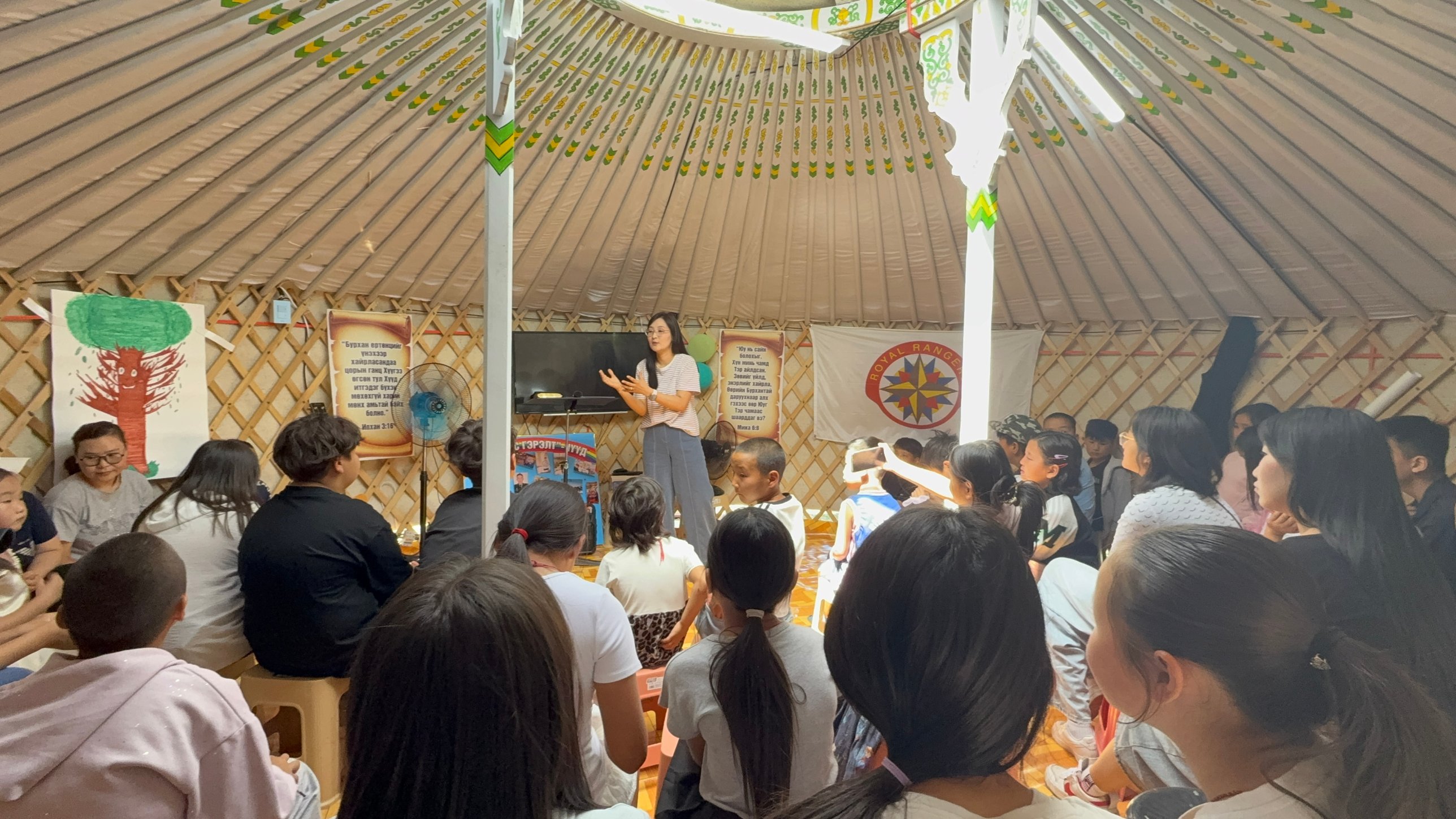 Mongolian children seated on the floor inside a traditional ger listening to a teacher speaking in front of a cross and scripture banners